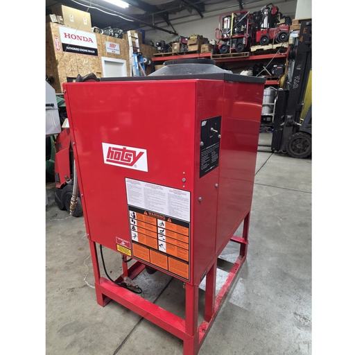 A red Hotsy industrial hot water heater with a metal frame, standing in a warehouse setting with other equipment and boxes in the background.